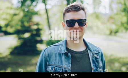 Close-up portrait of cheerful young man in sunglasses wearing trendy denim jacket looking at camera and smiling. Sunny summer day, beautiful green park in background. Stock Photo