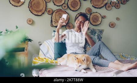 Happy African American teenager is making video call with smartphone calling friends talking and gesturing while her cute pet dog is lying on bed with her. Stock Photo