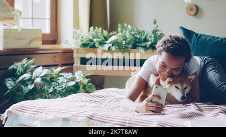 Joyful dog owner is taking selfie with cute friendly pet lying on bed together holding smartphone posing and hugging animal. Friendship between people and puppies concept. Stock Photo