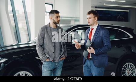 Handsome bearded guy is talking to car dealership manager in motor show discussing new automobile model, dealer is holding documents and pen. Purchasing vehicle and people concept. Stock Photo