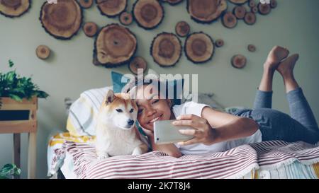 Cheerful girl is taking selfie with cute pet posing with beautiful dog lying on bed having fun and laughing. Modern apartment with lovely design and furniture is visible. Stock Photo