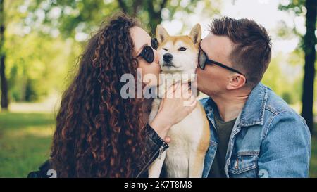 Proud dog owners pretty girl and handsome guy are playing with pet shiba inu dog, kissing it and scratching its fur relaxing in the park at weekend. People and animals concept. Stock Photo