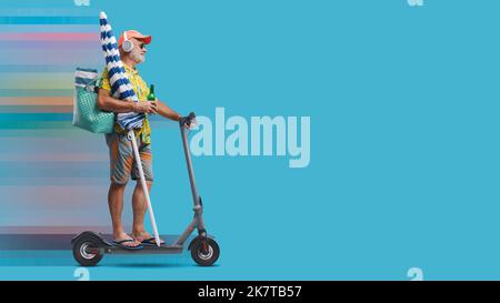 Funny happy senior tourist going to the beach, he is holding a beach umbrella and riding a fast electric scooter Stock Photo