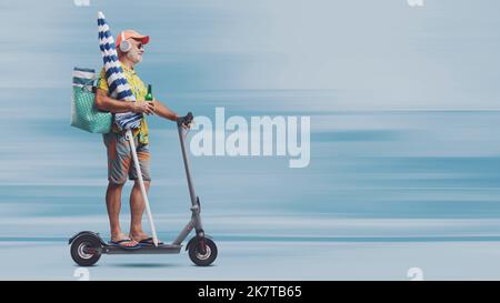 Funny happy senior tourist going to the beach, he is holding a beach umbrella and riding a fast electric scooter Stock Photo