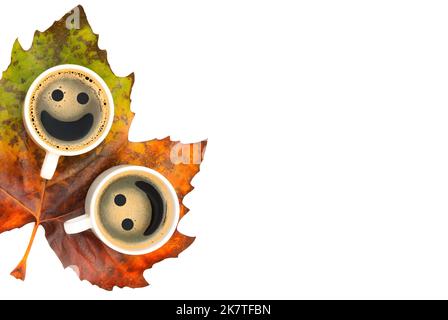 Two coffee cups with smiley faces placed on a large pigmented maple leaf isolated on white background with copy space. Stock Photo