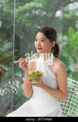 attractive asian smiling woman isolated on living room at home eating salad Stock Photo