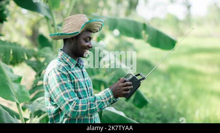 African farmer man listens retro radio broadcast receiver on shoulder stands happy smiling in his organic farm Stock Photo