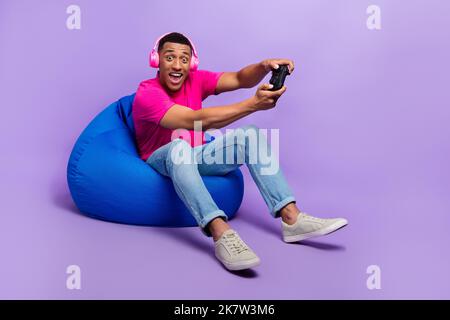 Photo of positive astonished man addicted user gamer wear stylish clothes sit comfort chair have fun isolated on purple color background Stock Photo