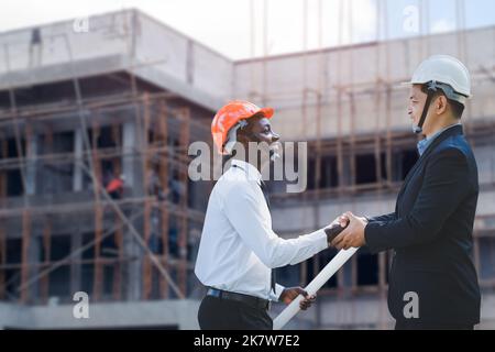 Smiling african engineer and asian construction managers shaking hands at construction site with happy at the bulding site. Concept of  team workers c Stock Photo