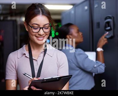 Regular maintenance is key. an attractive young female programmer working in a server room with her colleague in the background. Stock Photo