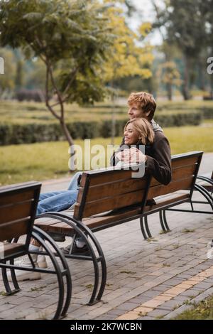 cheerful young man hugging woman and sitting on wooden bench in park,stock image Stock Photo