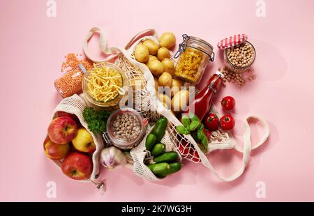 Top view of ripe vegetables and bottle of ketchup placed in string bags near glass jars of pasta and grains on pink background Stock Photo