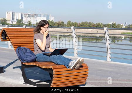 Girl student watching an online video lesson from a mobile phone on a bench. A teenager girl watches a video, listens to music in a smartphone with he Stock Photo