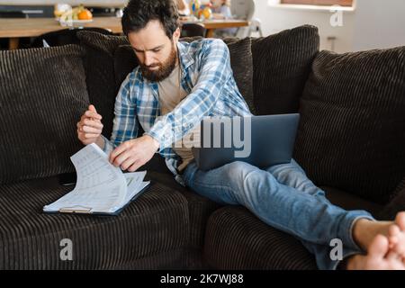 Middle aged brearded man working on laptop computer looking through documents sitting on a couch at home his wife and little son having breakfast at t Stock Photo