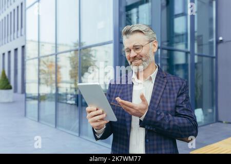 Senior gray-haired businessman outside office building, mature boss using tablet computer for remote communication video call with colleagues and employees, investor smiling and happy Stock Photo