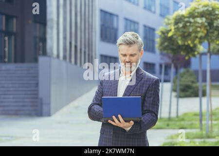 serious and concentrated gray haired boss working on laptop while standing outside modern office building, mature boss in business suit typing on netbook keyboard, senior successful businessman. Stock Photo
