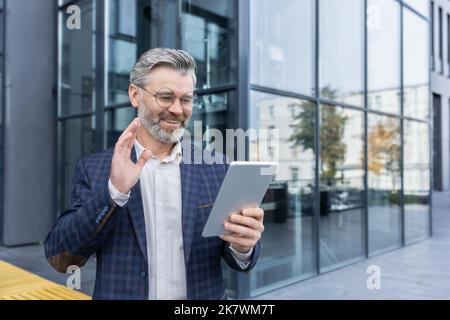 Senior gray-haired businessman outside office building, mature boss using tablet computer for remote communication video call with colleagues and employees, investor smiling and happy Stock Photo