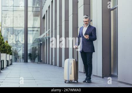 Business trip, senior gray-haired businessman outside hotel using phone, mature boss with big suitcase, investor in business suit and glasses. Stock Photo