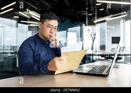 Thinking asian businessman working inside modern office building, man reading letter received from bank, boss thinking serious and concentrated working with laptop. Stock Photo