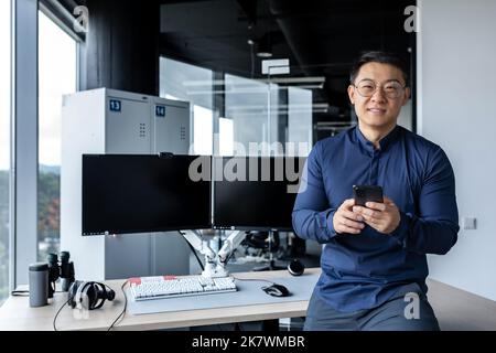 Portrait of successful businessman, man holding smartphone, asian man working inside modern office building using dual computer, worker looking at camera and smiling in glasses. Stock Photo