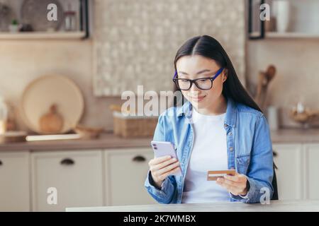 A young beautiful Asian woman in glasses and a denim shirt holds a phone and a credit card in her hands, uses. Sits at home in the kitchen, makes online purchases, makes an order, smilling. Stock Photo