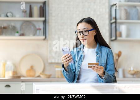 Financial problems. Worried young asian woman holding phone and credit card. Sitting in the kitchen at home, trying to solve a problem. Stock Photo