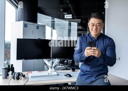Korean worker programmer developer working inside modern office building with dual computers, man using phone, typing message, Asian man reading good news online from smartphone. Stock Photo