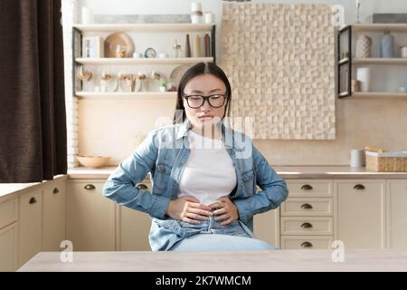 Young woman sick alone at home having severe stomach pain, asian woman holding her stomach with hands sad sitting on sofa in living room. Stock Photo