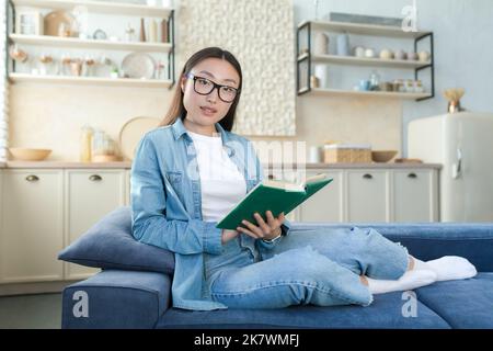 Portrait of a young beautiful Asian woman in denim clothes and glasses sitting at home on the sofa on the background of the kitchen. He holds a green book in his hands, reads, looks at the camera. Stock Photo