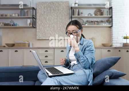 Work from home. Hospital. A young Asian woman freelancer fell ill. Works from home on the sofa at the laptop. He feels bad. He has a runny nose, wipes his nose with a napkin. Stock Photo