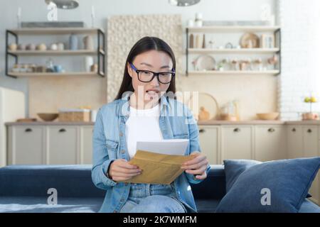 Happy asian girl shocked by received letter, woman reading message sitting on sofa at home. Stock Photo