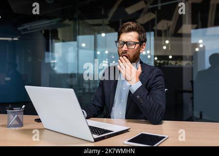 Mature bored businessman working inside modern office building, boss in glasses and business suit using laptop, investor yawning sitting on chair at table. Stock Photo