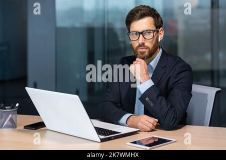 Serious thinking mature businessman working inside modern office building, senior boss in business suit and glasses working sitting at laptop, man with beard thinking about future strategy plan. Stock Photo