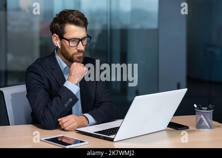 Serious thinking mature businessman working inside modern office building, senior boss in business suit and glasses working sitting at laptop, man with beard thinking about future strategy plan. Stock Photo