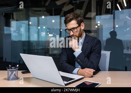 Serious thinking businessman working inside office building, man pondering important decision, boss in business suit using laptop at work. Stock Photo