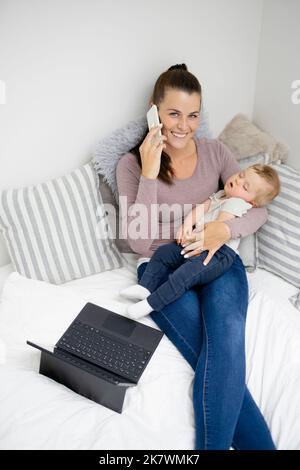 pretty young mother sitting, lying in bed, working on notebook and holding her sleeping baby in her arms and is happy Stock Photo