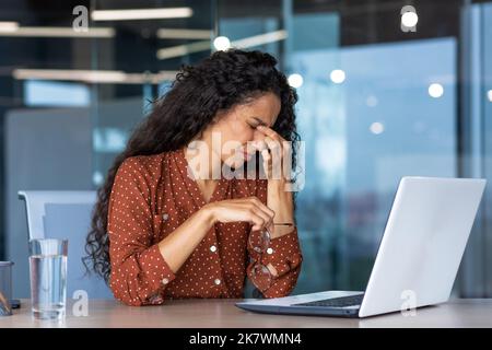 Tired business woman working inside modern office building, worker rubbing eyes with glasses off, latin american woman with curly hair using laptop for work. Stock Photo