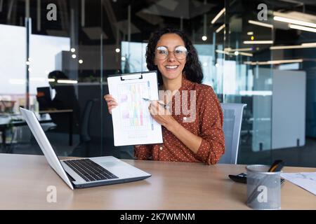 Portrait Hispanic business coach, woman smiling and looking at camera, showing documents with economic indicators to camera, teacher teaching remotely, mentor using laptop for online learning. Stock Photo