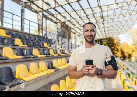 Sportsman using telephone smiling and looking at camera, man on morning jog and exercising at sports stadium, man listening to music in headphones and podcasts online radio. Stock Photo