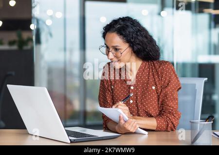 Young beautiful and successful business woman studying remotely sitting in the office, Hispanic woman writing in notebook using laptop for video call, talking with colleagues and mentor remotely Stock Photo