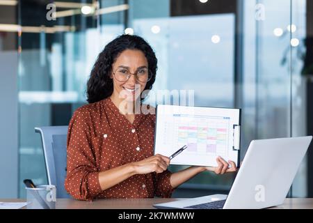 Portrait Hispanic business coach, woman smiling and looking at camera, showing documents with economic indicators to camera, teacher teaching remotely, mentor using laptop for online learning. Stock Photo