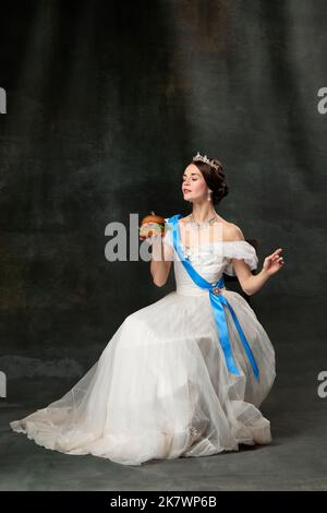 Refined taste. Young beautiful queen or princess in white medieval outfit eating burger on dark background. Concept of comparison of eras, art Stock Photo