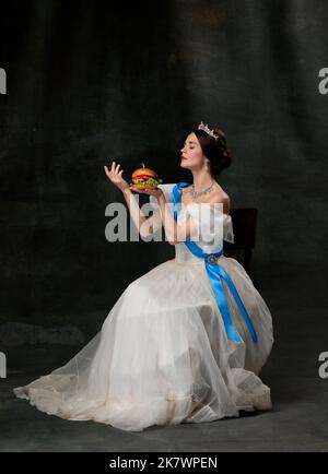 Refined taste. Young beautiful queen or princess in white medieval outfit eating burger on dark background. Concept of comparison of eras, art Stock Photo