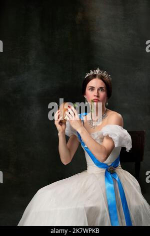 Fast food or dieting. Young funny girl like queen or princess in white medieval outfit eating burger on dark background. Concept of comparison of eras Stock Photo