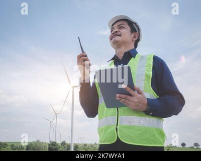 Asian engineer man stand holding tablet front the wind turbines generating electricity power station with talking by walkie -talkie . Concept of susta Stock Photo