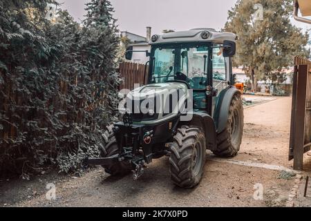 Modern green good-looking tractor stands in outside garage near agricultural fixtures and transport on farm or ranch on winter or summer cloudy day Stock Photo