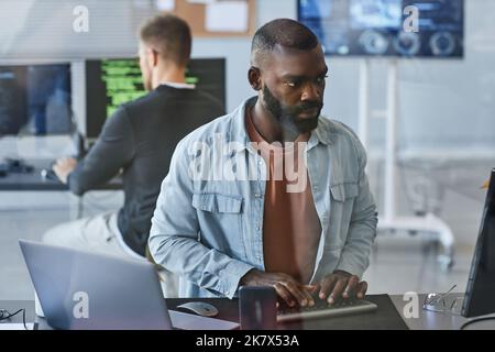Portrait of young black man using computer while programming software in IT company office behind glass wall Stock Photo