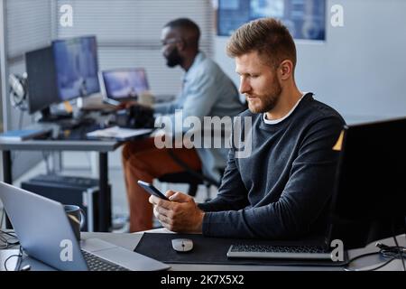 Side view portrait of young Caucasian man using smartphone at workplace while working in mobile software development, copy space Stock Photo