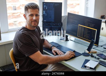 Portrait of young software engineer looking at camera while writing code at workplace with multiple computer screens Stock Photo