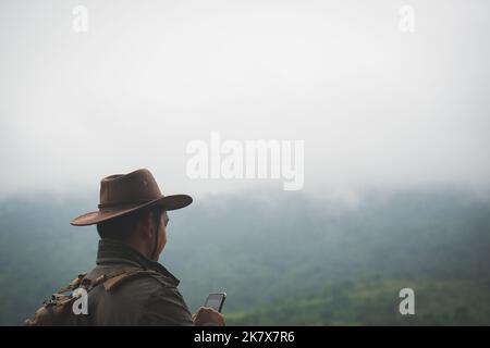 Freedom traveler man in hat carrying a backpack stands at the top of a mountain and using a smartphone on a foggy day.Adventure travel and success con Stock Photo